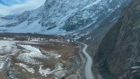 Vista-Aérea-De-Una-Carretera-Serpenteante-Que-Atraviesa-Un-Valle-Cubierto-De-Nieve-Rodeado-De-Escarpados-Picos-Montañosos.