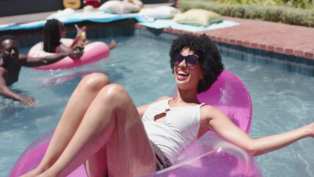 African American friends in swimwear at pool, man paddling spurring woman on pink ring laughing