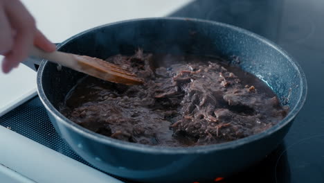 close up of a hand stirring the hot steaming beef meet with a wooden spoon cooking in a pan in slow motion during a day