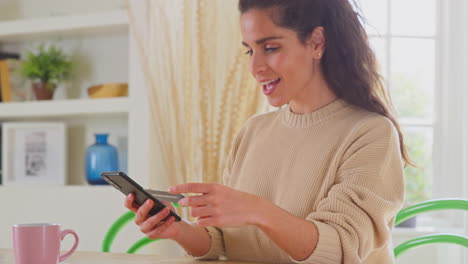 Celebrando-A-Una-Mujer-Con-Tarjeta-De-Crédito-En-Casa-Usando-Un-Teléfono-Móvil-Para-Realizar-La-Compra-De-Sus-Sueños