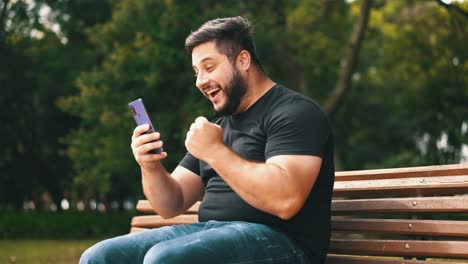 joven celebrando buenas noticias recibidas en su celular mientras está sentado en un banco del parque