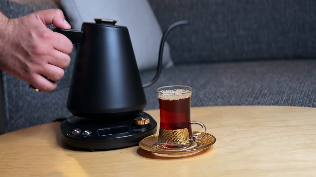 Close-up of hot tea being poured from a gooseneck kettle into a clear glass cup on saucer, electric kettle base beside it on a wooden table in a cozy indoor living room