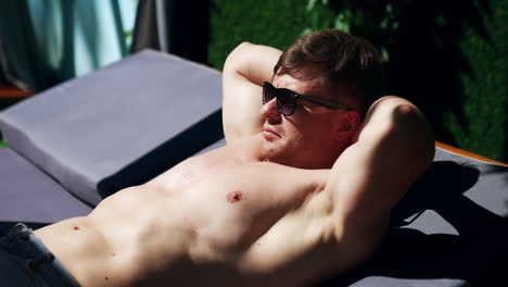 content topless caucasian man wearing sunglasses lies on the lounge. male holding hands behind his head and rises head to look around.