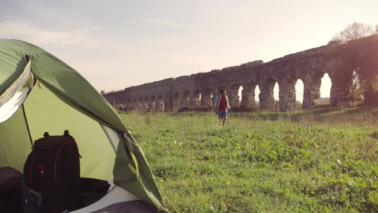 캠핑 텐트 밖에서 배을 고 있는 젊은 남자가 잔디 위에 지이를 들고 맨발로 걸어가며 로마 아쿠에듀크 앞에 서 있는 파르코 델리 아쿠에두티 공원 유적에 서서히 서서히 고 있다.