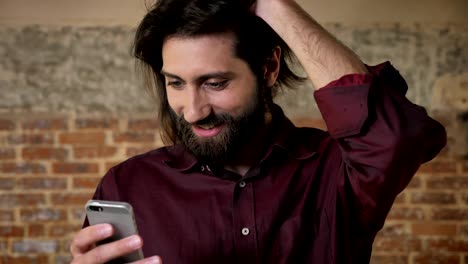 joven atractivo hombre moreno con barba está viendo en el teléfono inteligente, metiendo el cabello, fondo de ladrillo, concepto de comunicación