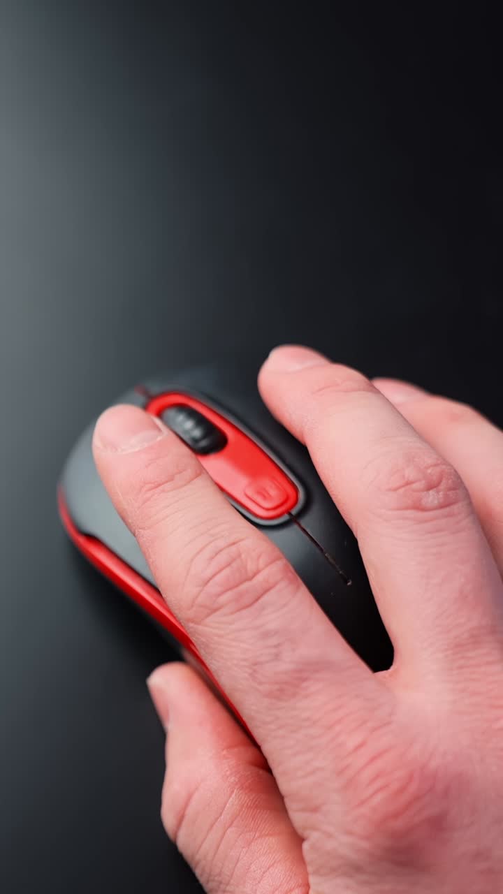 Close-up of a hand using a red and black computer mouse on a dark desk with copy space. Minimal tech vibe, soft light, focused productivity and calm mood