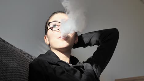 slow motion of young woman smoking electronic cigarette, releasing smoke in room with sunlight on her face