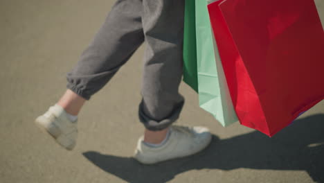 leg view of individual wearing grey joggers and white canvas shoes walking on tarred, carrying green, mint, and red shopping bags with a visible shadow cast on the ground