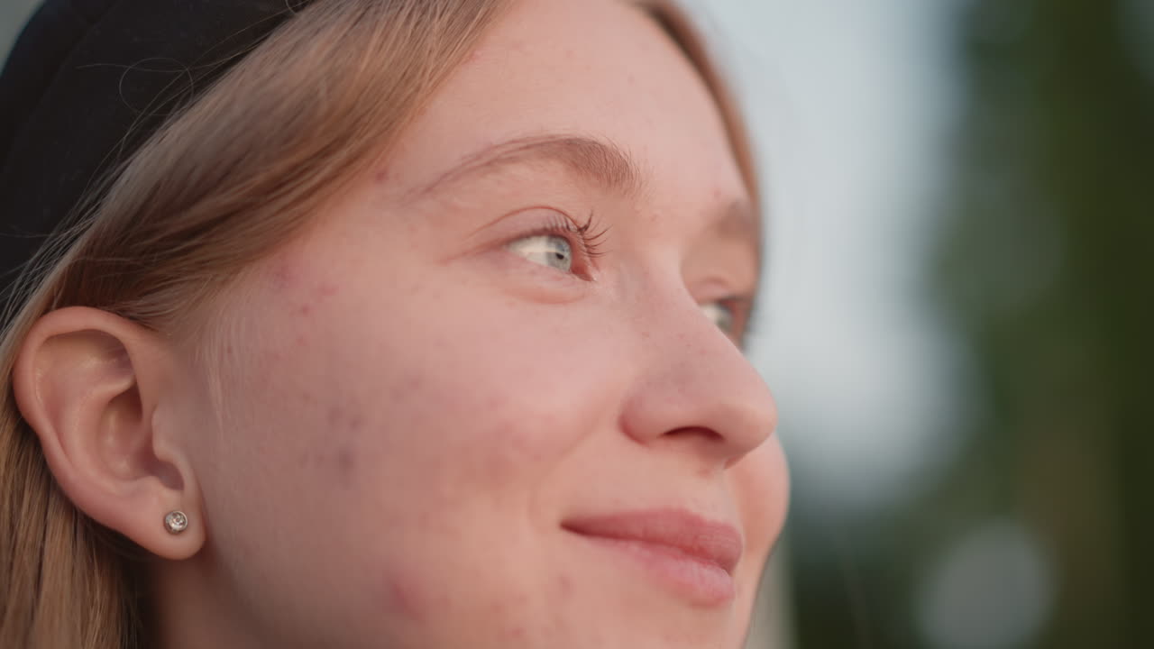 Primo piano di una giovane studentessa bianca che guarda in basso, con lentiggini e berretto, luce dell'ora d'oro e bokeh delicato, espressione calma, alberi sullo sfondo e un leggero sorriso speranzoso