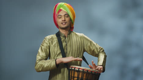Portrait-of-young-cheerful-Indian-man-in-traditional-clothes-and-turban-playing-a-drummer-and-smiling-at-camera