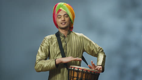 portrait of young cheerful indian man in traditional clothes and turban playing a drummer and smiling at camera