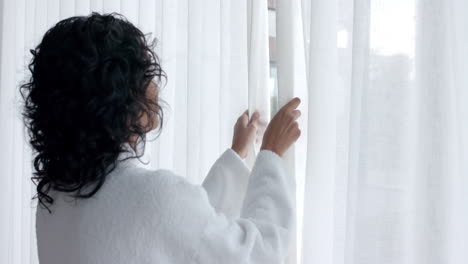 mujer biracial con bata de baño abriendo cortinas y mirando por la ventana en casa, cámara lenta