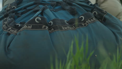 close up of blue duffle bag with grommet straps sliding across green grass as unseen teammates drag equipment toward paramotor launch site during calm dusk