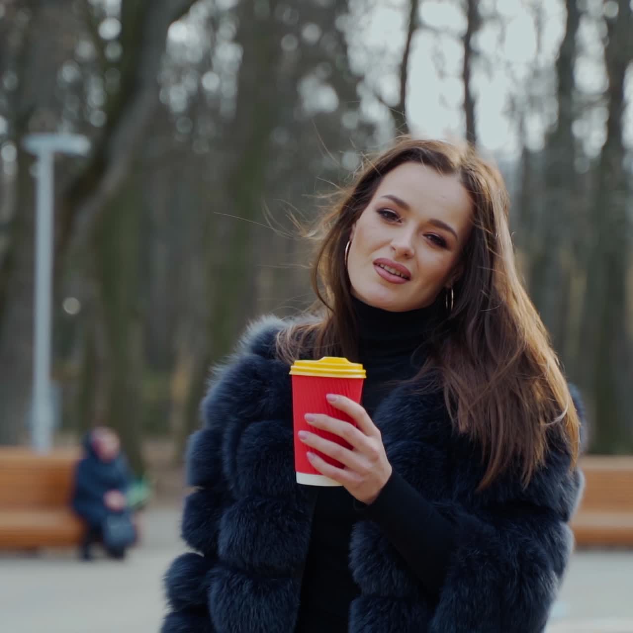 Young woman in fur coat in the park. Back view of a girl turning to camera with a plastic cup of coffee. Portrait of a stylish woman outdoors.