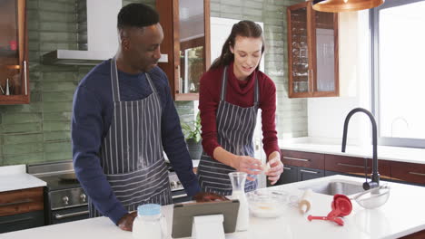 happy diverse couple standing in kitchen, using tablet and preparing dough,slow motion