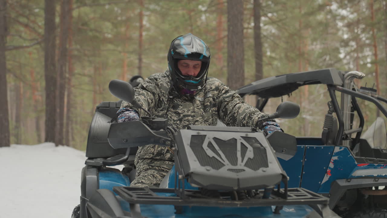 Uomo caucasico avvia un ATV sulla neve, un pilota adrenalinico si assicura casco e occhiali, accende l'UTV blu, afferra l'acceleratore, si tuffa in un sentiero di pineta, rombo del motore e fiato appannato, una corsa invernale estrema.