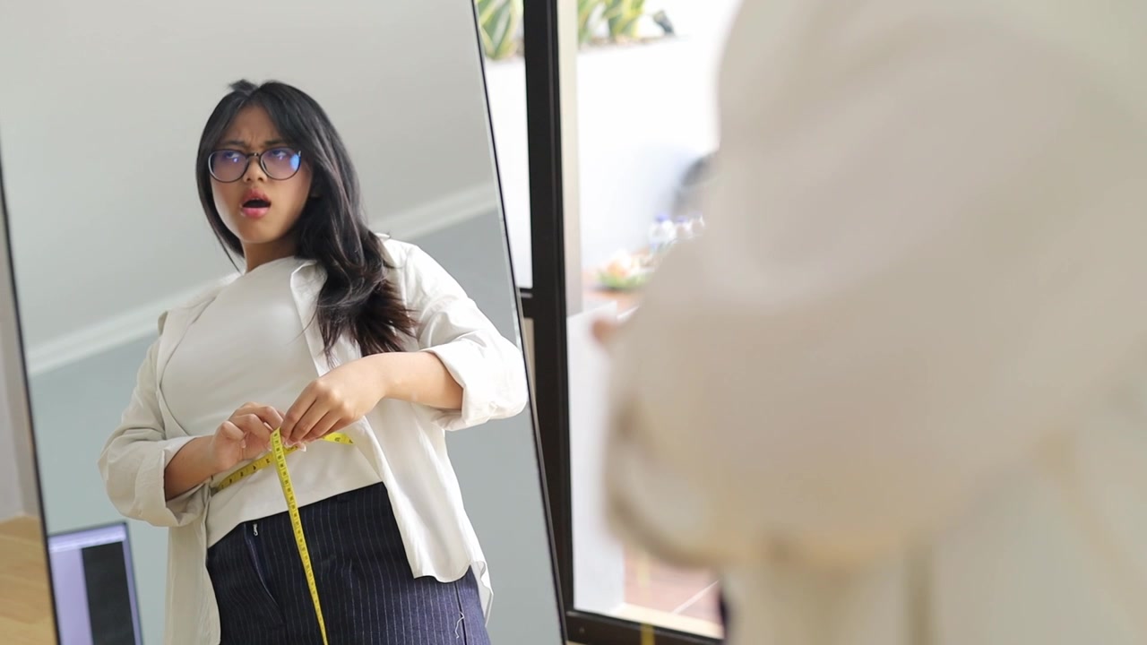 Unhappy Young Asian woman measuring waist near mirror, upset with visible weight gain