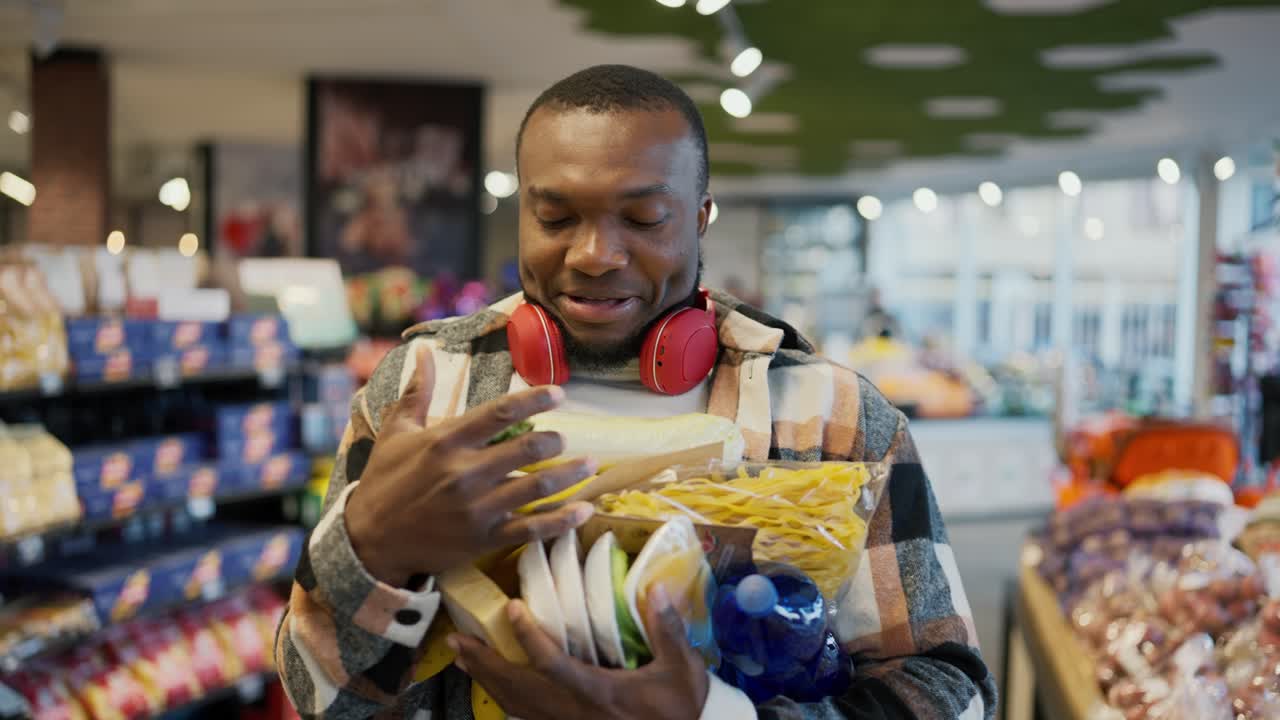 A happy and smiling man with Black skin in a checkered shirt and red wireless headphones carries a bunch of groceries on his chest holding with both hands during his walk and shopping in a modern supermarket