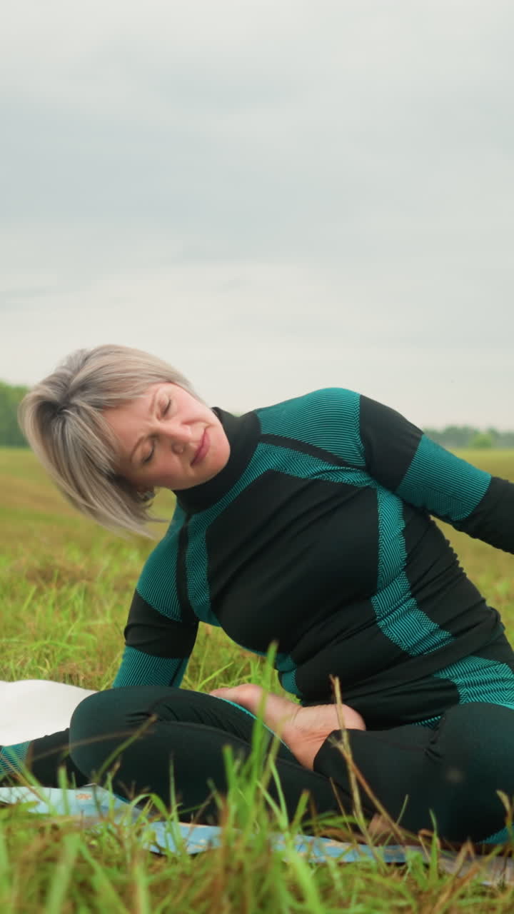 Woman with grey hair seated on yoga mat practicing side bend pose with hands stretched in a vast grassy field under cloudy skies, surrounded by nature with trees in the distance