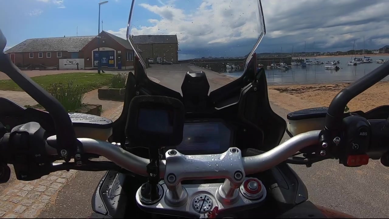 Motociclista in moto verso un porto in Scozia con cieli azzurri e yacht nel mare