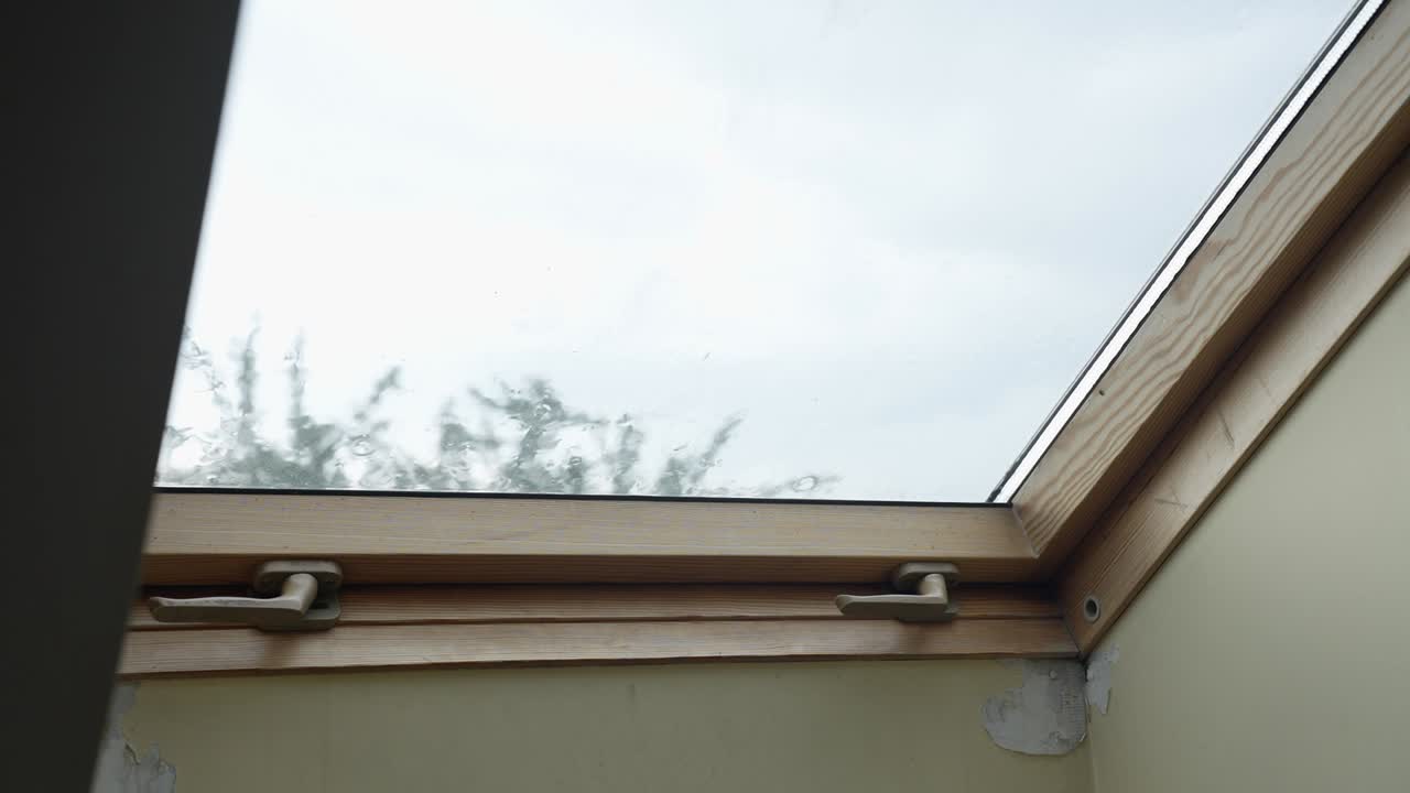 Static shot capturing raindrops landing on a slightly opened skylight window in an attic room, with soft light filtering through the glass and a blurred view of treetops in the background