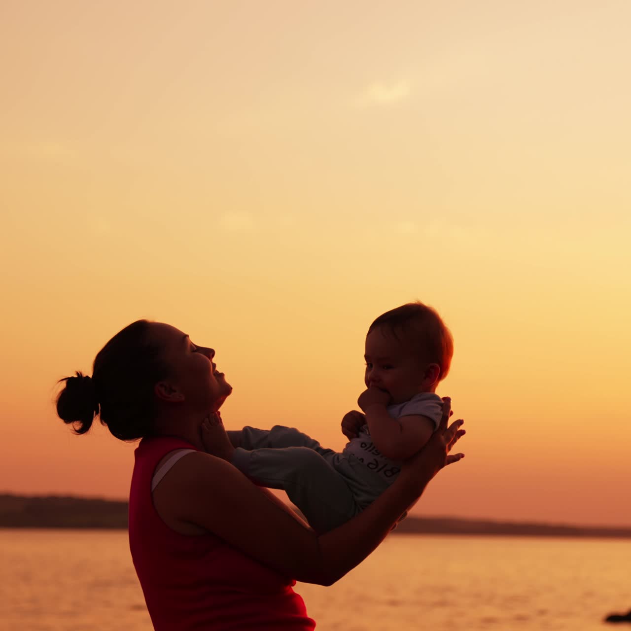 Happy mom playing with her little baby near the river at sunset. Woman rises her son up in the air and kid put little feet on her face