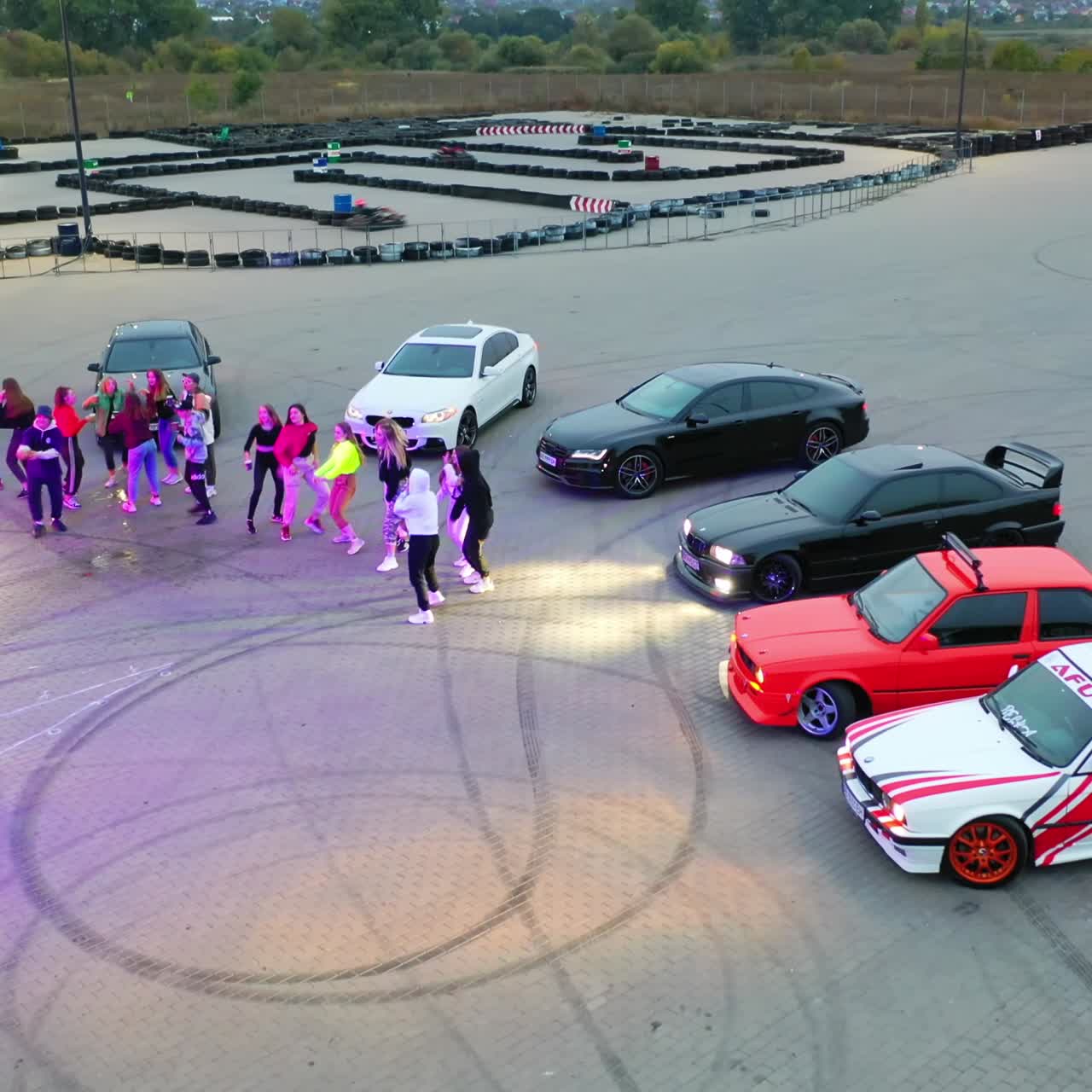 Party at the drifting field. Young happy people dancing near the race cars outdoors at dusk. Top view