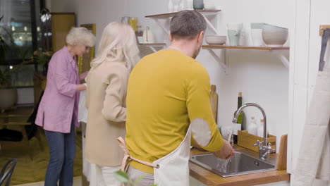 vista trasera de un hombre lavando los platos de la cena familiar en el fregadero de la cocina mientras dos mujeres maduras quitan los platos de la mesa