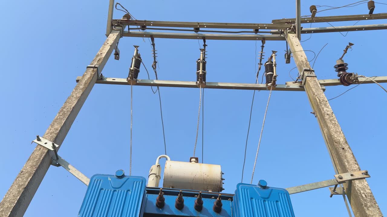 Video con ripresa dal basso verso l'alto di un trasformatore di distribuzione elettrica montato su un palo della luce, con cavi isolati e bracci a crociera visibili su un cielo azzurro.