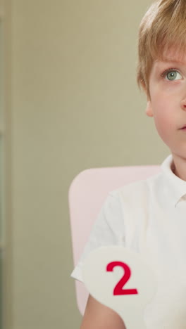 diligent toddler boy raises tag with digit sitting at desk at maths lesson. cute preschooler child learns numbers and solves tasks in nursery classroom