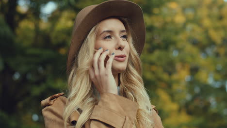 Close-up-view-of-caucasian-young-blonde-woman-wearing-a-hat-and-talking-on-the-phone-in-the-park-in-autumn