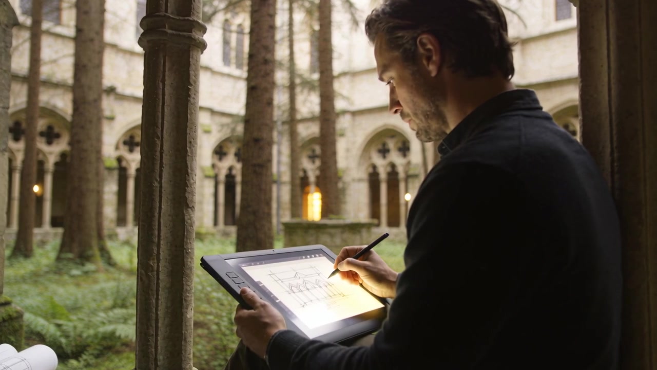 En mand i jakke skitserer planer på en tablet i et kloster, kameraet kører tættere på, med plads til tekst.