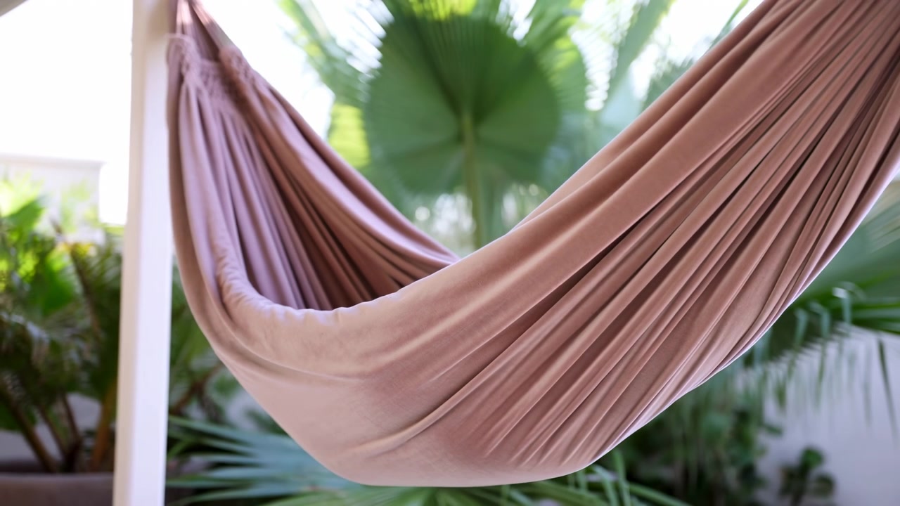 Movimento ravvicinato di un'amaca in tessuto malva in una veranda tropicale, che cambia lentamente da una tasca curva e rilassata a una forma leggermente tesa e pieghettata sotto la luce del giorno, con punti luce morbidi, toni smorzati e ambiente interno.