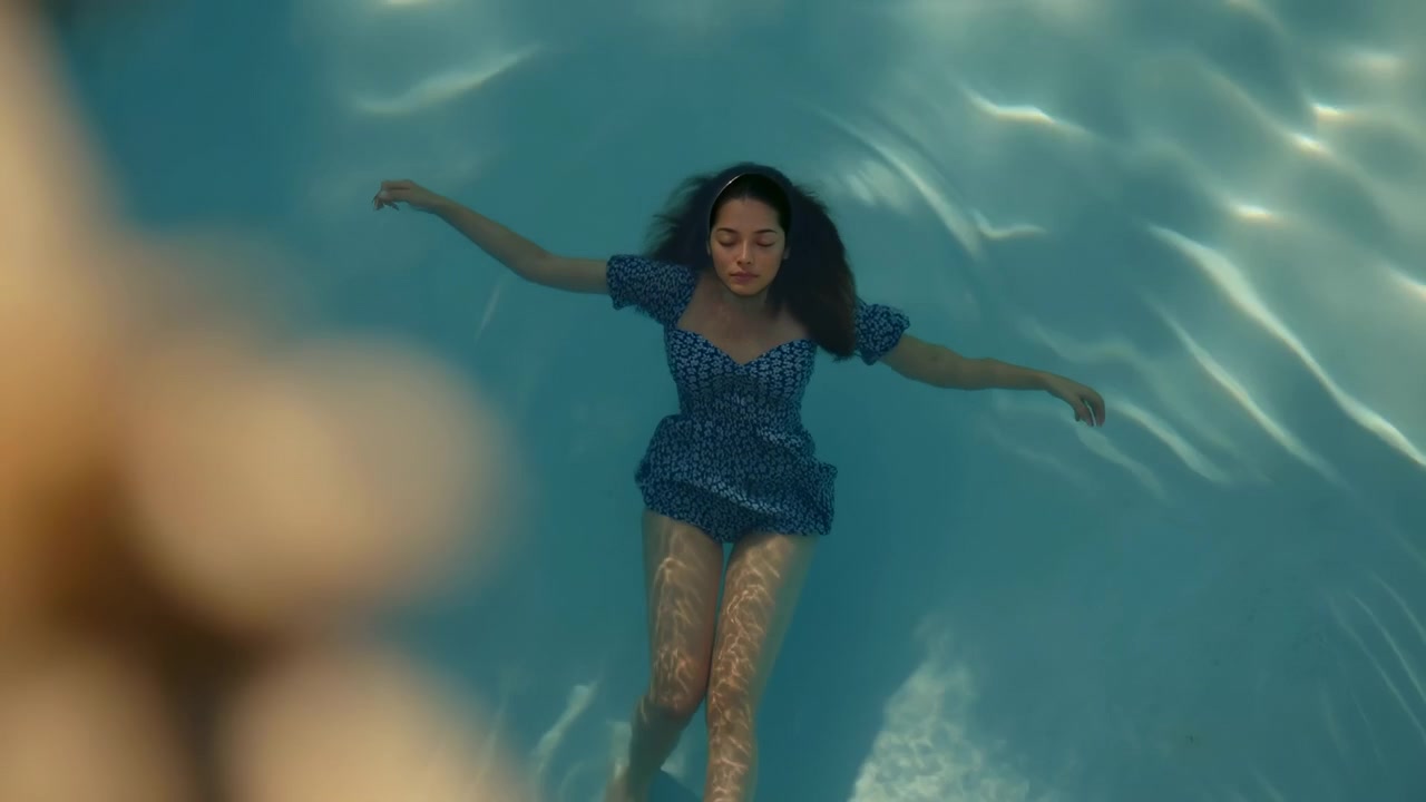 Top-down view shows her floating almost motionless below the surface, while refracted light shimmers over her legs and pale blue floor and her dark hair fans outward.