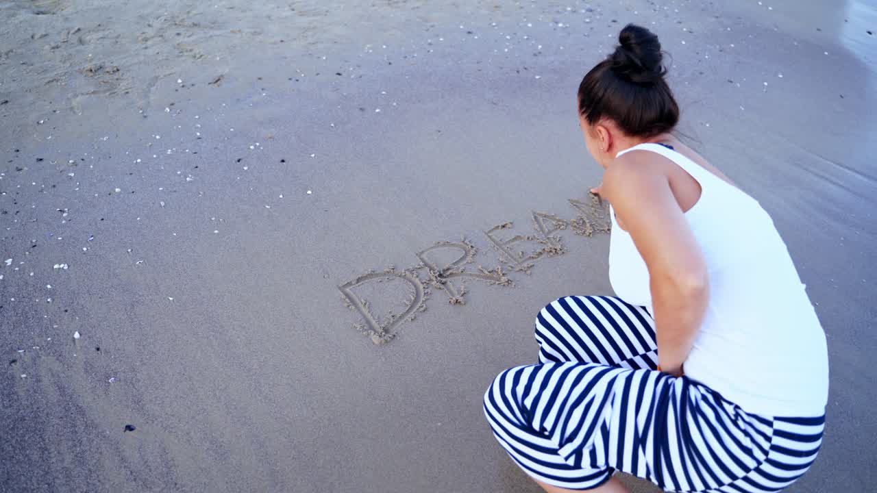 Back view of a female on the shore.Woman writing the word dream on a sandy beach near the sea. Water waves washing off the written word. Top view.