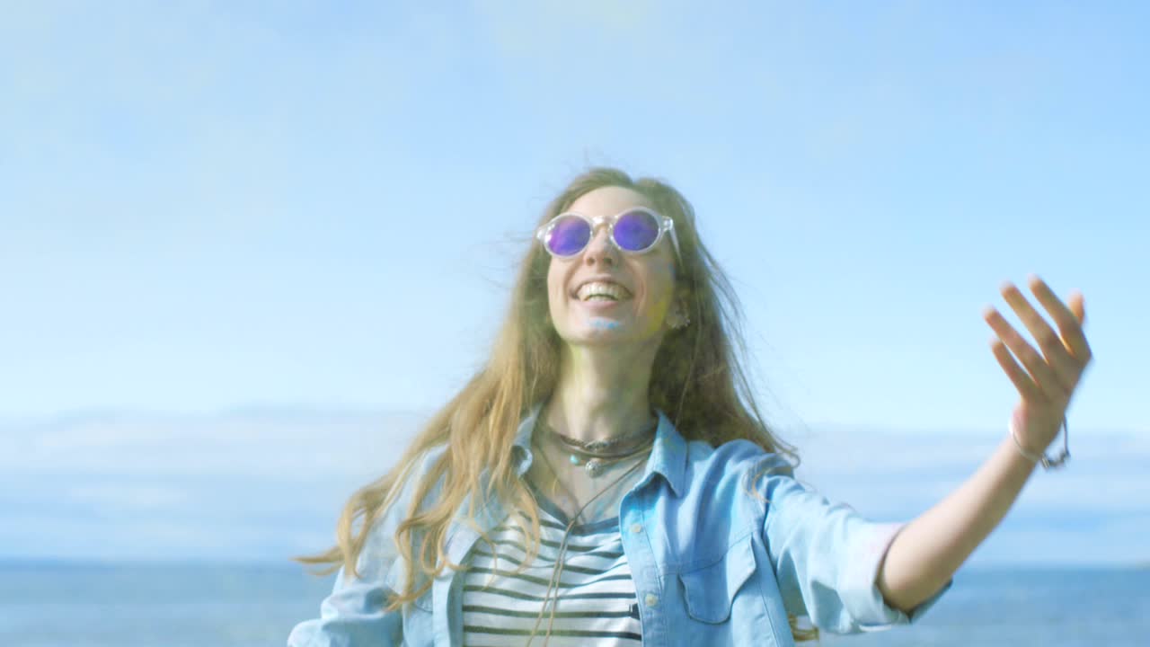 Beautiful Young Young Girl with Brown Hair Wearing Cool Sunglasses Throws Holi Colorful Powder Into the Air and Laughs. Clear Blue Sky Behind Her and Deep Blue Sea Behind Her.
