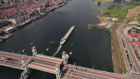 Elevated-bridge-over-a-river-in-a-european-town-with-boat-passing-under,-cars-and-cyclists-waiting,-cloudy-day,-slight-haze,-aerial-view