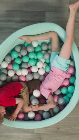 positive toddler boy and preschooler girl crawl among colorful plastic balls in elastic dry pool. playful couple of kids plays together at home