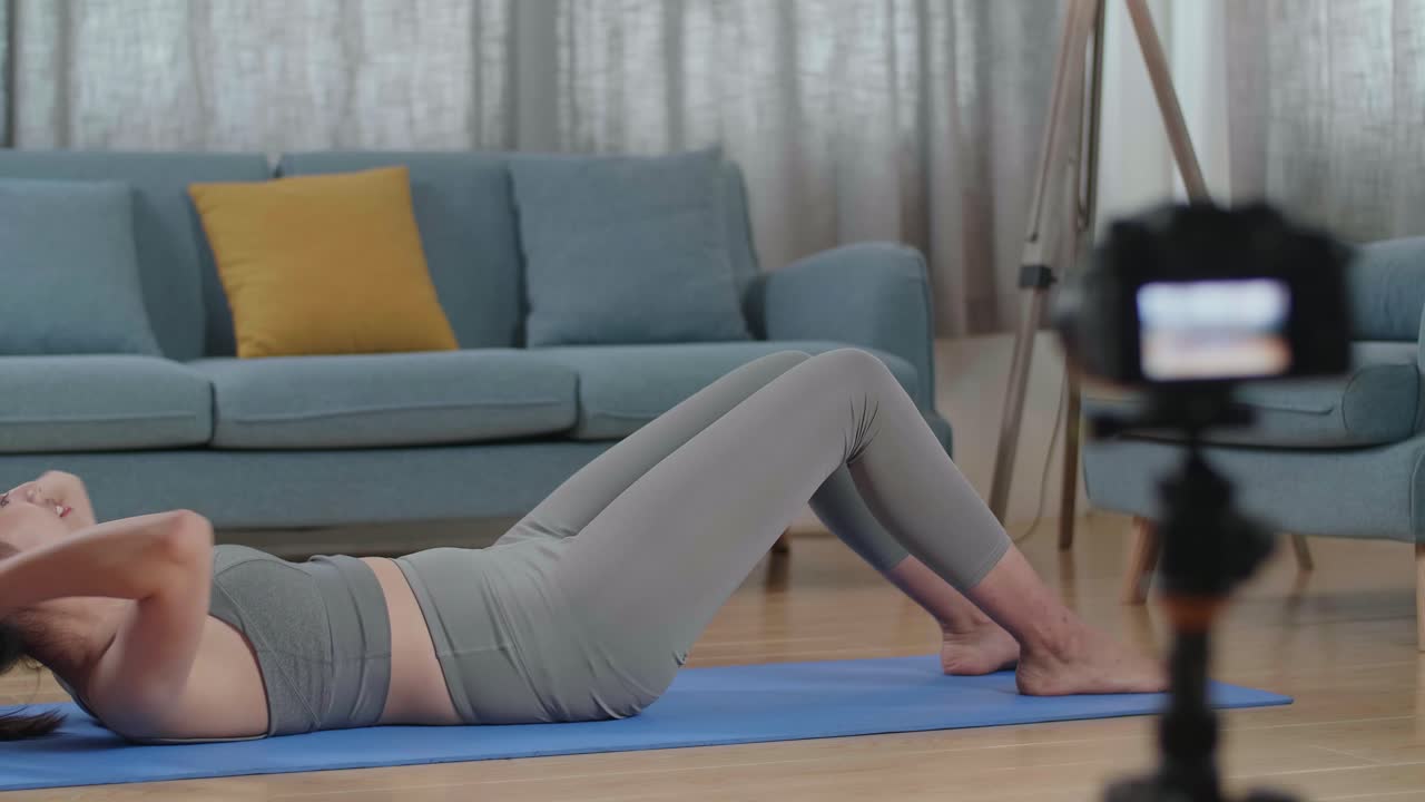 joven entrenadora asiática mujer en ropa deportiva hablando a la cámara y haciendo ejercicios de crisscross crunch mientras graba ejercicios de enseñanza en casa