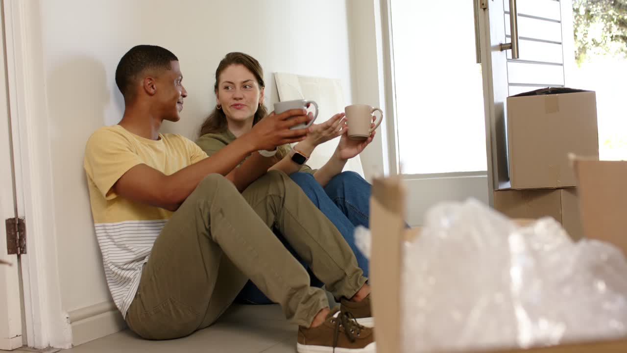 Pareja diversa sentada en el suelo junto a cajas de mudanza, haciendo un descanso mientras desempaquetan y bebiendo de tazas de café
