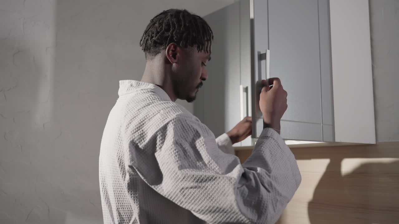 Close-up of man in bathrobe with short hair, facing sideways. Strong lighting emphasizes his profile, with shadows on the wall. Minimalist background adds depth and focus on his features