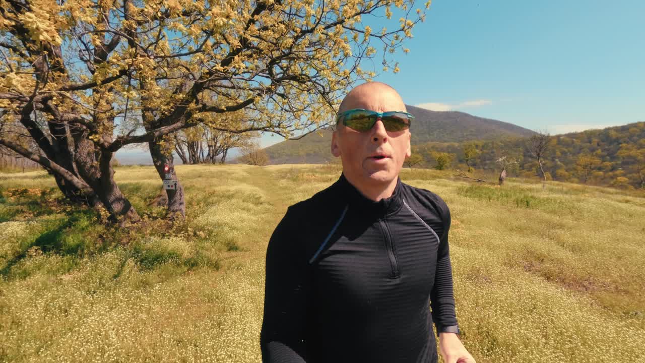 Active mature man in his 50s running on a trail through a scenic field and forest on a sunny spring day. Healthy senior lifestyle, jogging and exercising outdoors. High quality 4k footage
