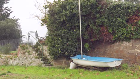 a small blue and white sailboat rests on the grass beside a stone wall overgrown with ivy. the atmosphere is calm and wintry, in a quiet coastal garden setting.