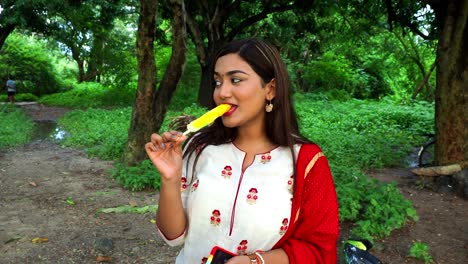 Beautiful-young-woman-standing-on-lake-and-eats-ice-cream-in-stick-with-tress-and-grass-in-the-background