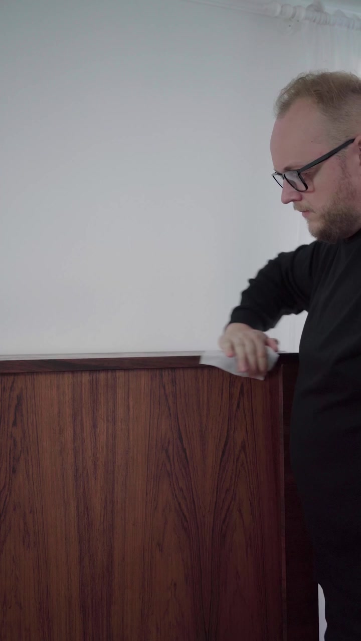 Uomo con maglione nero lucida un mobile in teak vintage con un panno, rifinendo la finitura del legno trattato in un interno luminoso.