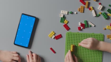 Overhead-Shot-Of-Two-Children-Playing-With-Plastic-Construction-Bricks-Next-To-Blue-Screen-Mobile-Phone