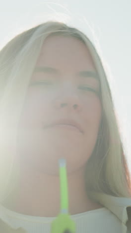 primer plano de una mujer joven de pie bajo la brillante luz del sol, sosteniendo una bomba de aire verde para enfriar su cara, con su cabello rubio revoloteando suavemente en la brisa, su expresión es tranquila y enfocada