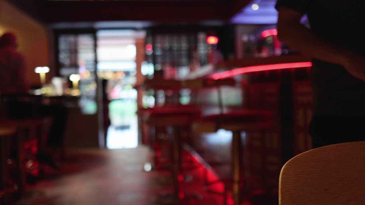 A server walks out of a dimly lit, modern bar and restaurant with red neon accents, captured in a smooth, stationary wide shot