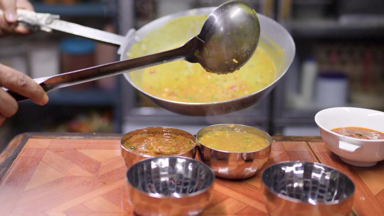 Primo piano di uno chef che versa curry caldo di lenticchie gialle in ciotole di metallo in una cucina commerciale.