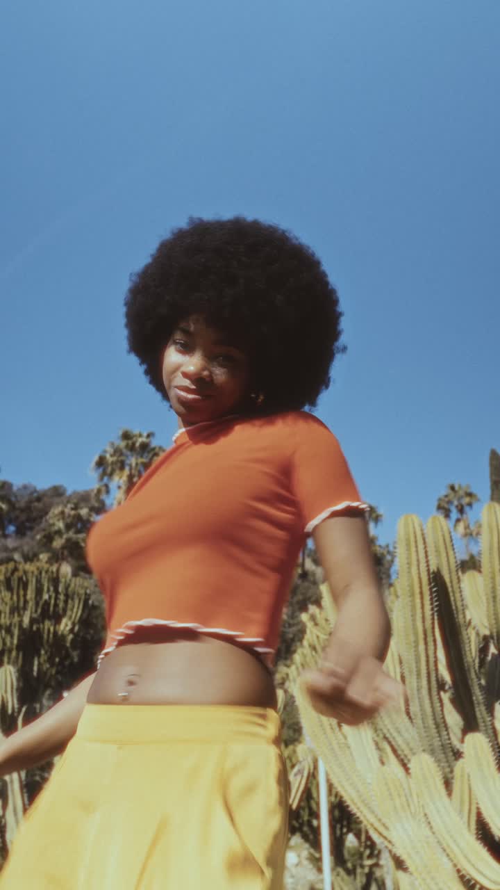 Giovane donna vivace con capelli afro che balla tra i cactus sotto il cielo azzurro